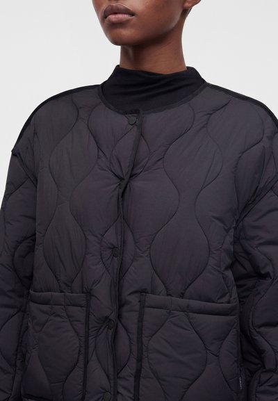 Personne portant une veste matelassée noire avec un col rond et des boutons-pression sur le devant, dotée de deux grandes poches avant, montrée des épaules vers le haut.