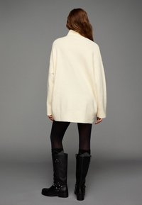 Överdimensionerad krämfärgad tröja med hög krage, matchad med svarta knähöga stövlar. Texturerat tyg. Mörka leggings kompletterar outfiten.