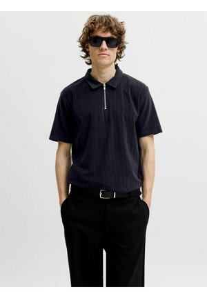 Jeune homme aux cheveux bouclés portant des lunettes de soleil noires, un polo sombre à manches courtes avec une fermeture éclair, et un pantalon noir les mains dans les poches.