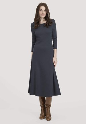 Woman with long brown hair wearing a fitted navy midi dress with three-quarter sleeves and brown knee-high boots, standing against a plain background.
