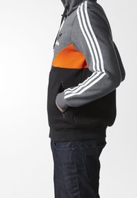 Sudadera con capucha en paneles grises, naranjas y negros. Presenta tres franjas blancas en las mangas y un bolsillo con cremallera en la parte frontal.