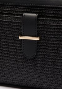 Schwarze gewebte Tasche mit einer strukturierten Oberfläche. Verfügt über einen glatten Lederträger, der mit einer goldenen Metallbalkenschließe gesichert ist.