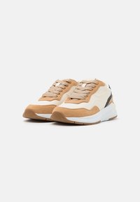 Füllige Sneakers in Beige mit braunen Wildlederelementen, ausgestattet mit texturiertem Stoff, runder Zehenpartie und flacher Gummisohle für zusätzliche Stabilität.