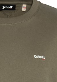 Olivovo zelené tričko vyrobené z bavlny s okrúhlym výstrihom, na prednej strane s malým bielym logom "Schott" a s vnútornou etiketou s vlajkou.