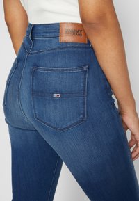 Denim jeans in dark blue with a high waist, featuring a leather Tommy Jeans patch, and two back pockets with a distinct logo detail.