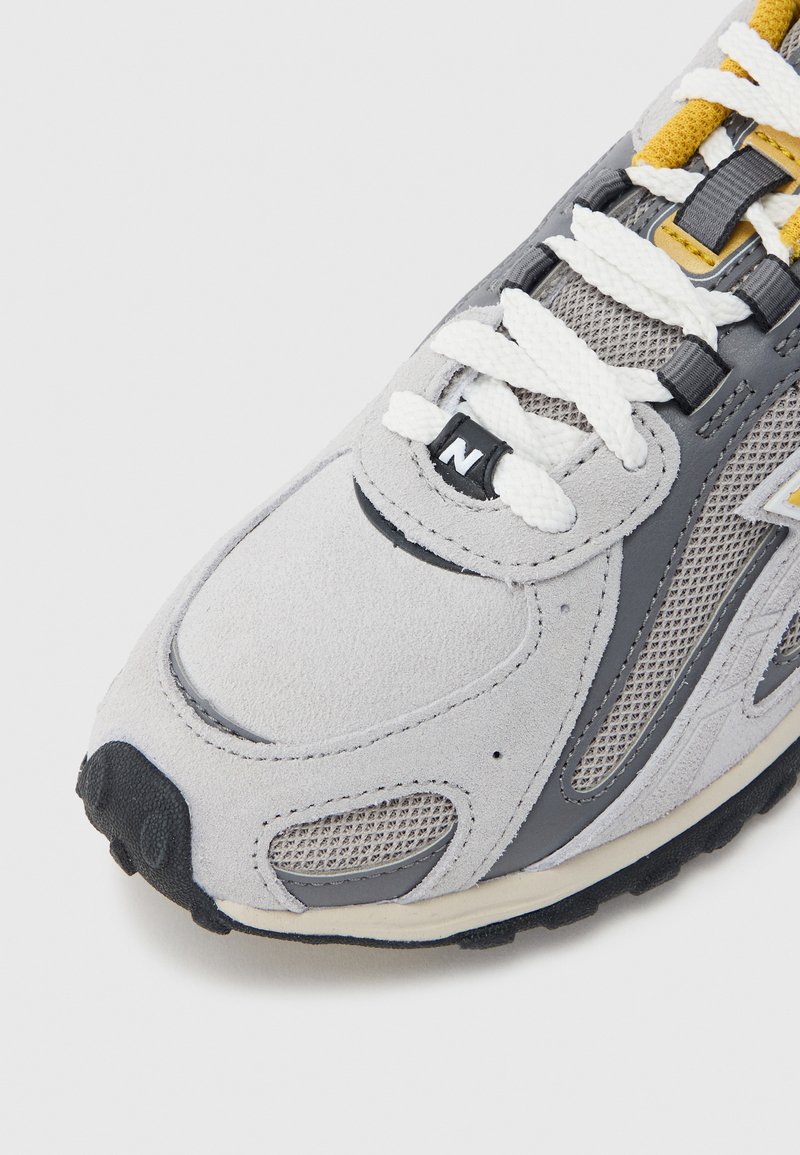 Grauer Laufschuh aus Wildleder und Mesh mit weißen Schnürsenkeln, gelben Akzenten und schwarzer Gummisohle. Auffälliges Logo und atmungsaktive Designdetails.