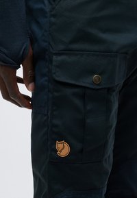 Mørkeblå bukser med klaffelomme sikret med en messingknapp og et brunt reverlogo-merke nær lommen på låret.