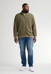 Olivengrön zip-up fleecejacka med texturerat tyg, hög krage och sidofickor, matchad med ljusblå jeans och vita sneakers.