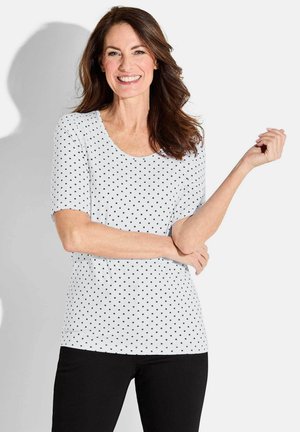 Witte mouwloze top met een ronde hals, versierd met een patroon van zwarte stippen. Draagt met zwarte broek. Gladde textuur.