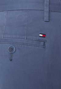 Blå bomullsbyxor med en bakficka som har knappstängning, detaljerade med en liten röd, vit och blå broderad logotyp nära fickan.