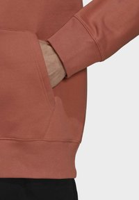 Person som bär en rustfärgad sweatshirt med en hand placerad i den främre kängurufickan framför en enfärgad bakgrund.