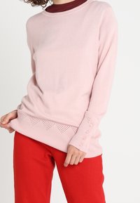 Jasnoróżowy sweter z długim rękawem z ribbowanym dołem, ozdobiony zygzakowatym wzorem oraz guzikami na rękawach, noszony na maroon topie.