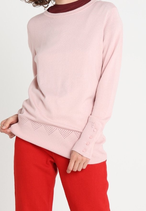 Jasnoróżowy sweter z długim rękawem z ribbowanym dołem, ozdobiony zygzakowatym wzorem oraz guzikami na rękawach, noszony na maroon topie.