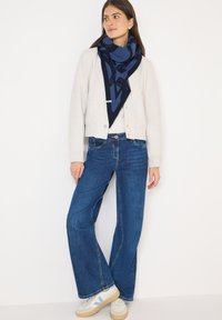 Marineblåt mønstret tørklæde lagdelt over en lysegrå cardigan, kombineret med mørkeblå flared jeans og cremede sneakers med lyseblå accenter.