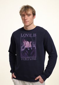 Giovane uomo che indossa una felpa blu navy con una grafica viola di una ragazza con trecce e il testo "L'amore è tortura." Mani nelle tasche.
