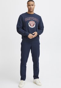 Tengerészkék pulóver "HARVARD" felirattal és emblémával a mellrészen, tengerészkék nadrággal és fehér sportcipővel párosítva. Simább anyagú, laza szabású.