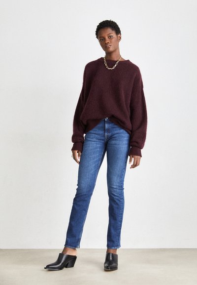 Suéter burdeos de gran tamaño con tejido texturizado, combinado con jeans de mezclilla azul de corte ajustado y mule de tacón abierto en negro. Collar de cadena dorada.