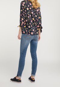Blusa negra con estampado floral, de mangas tres cuartos y detalles de lazo, combinada con jeans ajustados azules y zapatillas negras.