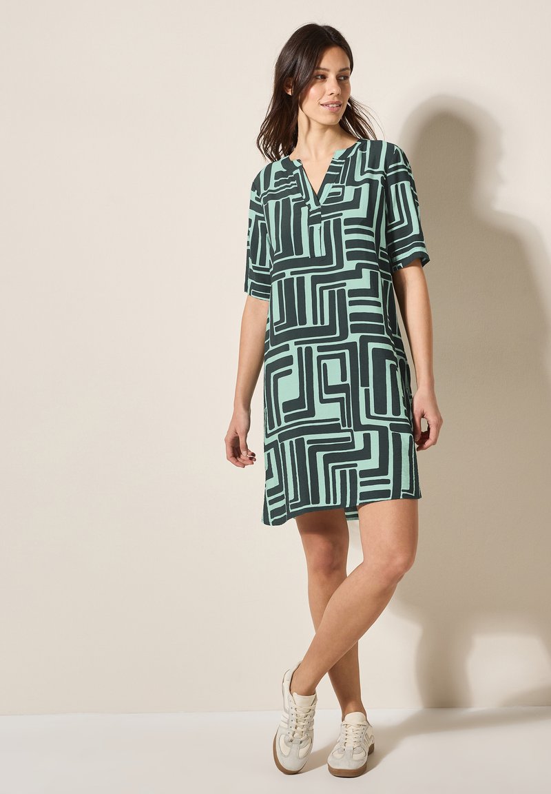 Grünes gemustertes Kleid mit schwarzen geometrischen Linien, kurzen Ärmeln und einem V-Ausschnitt. Kombiniert mit weißen Sneakers. Glatte Stofftextur.