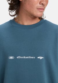 Sudadera color verde azulado de algodón, con cuello redondo, textura suave, y logo blanco con estampado gráfico centrado en el frente.
