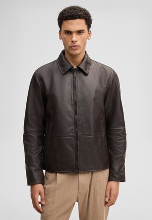 Homme aux cheveux courts et bouclés porte une veste en cuir noire zippée et un pantalon beige clair, debout devant un fond uni gris clair.