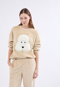 Beżowa bluza polarowa z pluszowym wzorem pudla. Posiada luźne rękawy, okrągły dekolt oraz pasujące spodnie dresowe. Miękka tekstura.