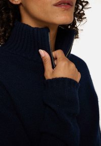 Pull bleu marine avec un col côtelé haut et une fermeture éclair complète. Présente un tissu tricoté texturé et des poignets côtelés, mettant en avant la chaleur et le confort.