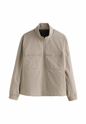 Giacca beige con zip e colletto alto, due tasche con zip sul petto, maniche lunghe e polsini a coste, esposta su sfondo bianco.