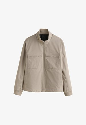 Bežs, ar rāvējslēdzēju aizdaram, džemperis ar stāvu apkakli, divām krūšu kabatām ar rāvējslēdzēju, garām piedurknēm un aprocēm, novietots uz baltas fona.