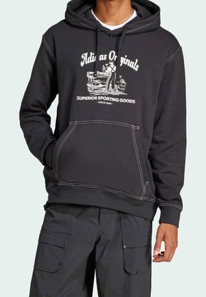 Sweat à capuche noir avec poche avant, imprimé blanc « Adidas Originals » et graphique d'ouvrier, porté avec un pantalon cargo noir, porté par une personne debout.