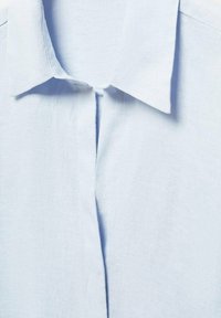 Chemise en lin bleu clair avec un col classique, une patte de boutonnage et une texture lisse, mettant en valeur de subtiles rides et des détails de couture.