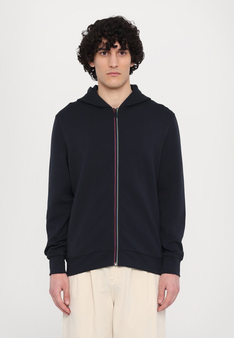 Sudadera con cremallera en color azul marino, con capucha, mangas largas y rayas multicolores en la cremallera. Presenta una textura suave y un corte relajado.