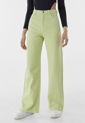 Kobieta ubrana w jasnozielone szerokie spodnie z wysokim stanem, białe trampki oraz czarną bluzkę z długim rękawem, skróconą na wysokości talii.