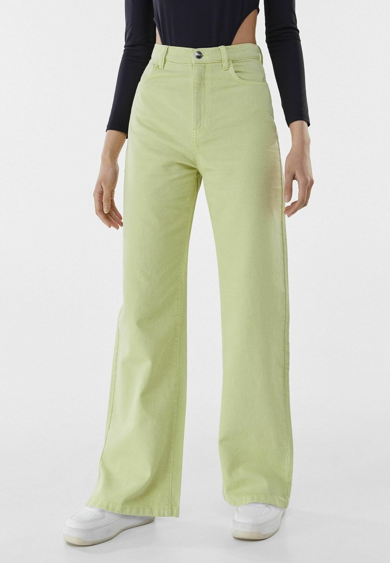 Femme portant un pantalon taille haute ample couleur vert clair, des baskets blanches et un haut noir à manches longues raccourci à la taille.