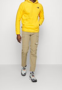 Gul huva sweatshirt med en framficka och logotyp, beige cargobyxor med sidofickor, och grå sneakers med orangea detaljer.