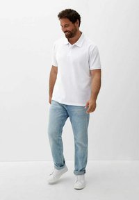 Polo shirt blanc à manches courtes avec col, associé à un jean bleu clair et des baskets blanches. Le modèle se tient sur un fond uni.