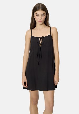 Mujer con cabello largo y castaño, vestida con un vestido negro sin mangas con tirantes finos y detalles de lazo en la parte delantera, posando frente a un fondo liso.