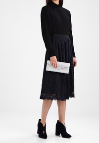 Czarna bluzka z długim rękawem, granatowa koronkowa spódnica midi, czarne botki do kostek z wycięciami, trzymająca srebrną kopertówkę.