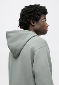 Ανοιχτό γκρι φούτερ με απαλό ύφασμα, διαθέτει κουκούλα με κορδόνια και άνετη εφαρμογή. Οι ραφές είναι ορατές κατά μήκος των ώμων και των μανικιών.