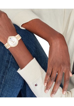 Montre analogique dorée avec un cadran rond, un cadran blanc et des accents argentés. Portée au poignet contre un jean foncé et le poignet d'un pull crème.