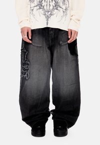 Χαλαρά μαύρα ξεθωριασμένα τζιν με φθαρμένα σημεία στους μηρούς, σε συνδυασμό με μαύρα αθλητικά παπούτσια και μακρυμάνικο πουκάμισο με σκούρο γραφικό σχέδιο.