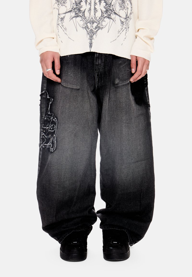 Χαλαρά μαύρα ξεθωριασμένα τζιν με φθαρμένα σημεία στους μηρούς, σε συνδυασμό με μαύρα αθλητικά παπούτσια και μακρυμάνικο πουκάμισο με σκούρο γραφικό σχέδιο.