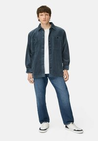 Mand iført en mørkeblå fløjlsovertrøje, hvid t-shirt, blå jeans og hvide sneakers, stående mod hvid baggrund.