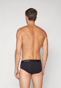 Fekete pamut alsónadrág puha derékkal, amelyen fehér "Calvin Klein" logó található; sima textura és kényelmes viselet. Háttal nézve.