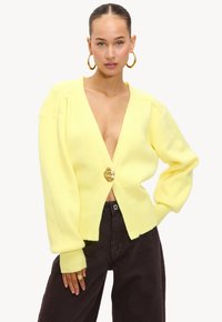 Geel gebreide cardigan met een diepe V-hals en een grote gouden knoop. Heeft ruime mouwen en een geribbelde textuur aan de onderkant en de manchetten.