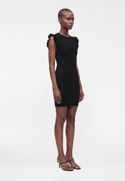 Femme portant une robe noire cintrée sans manches avec des épaules à volants et des sandales à talons noires à brides, debout devant un fond uni.