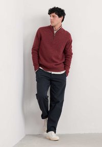 Sarkans zip-kakla džemperis ar teksturētu apdari, saskaņots ar tumšiem sašaurinātiem biksēm un baltiem sporta apaviem. Adīta auduma, ribota piedurkņu un apakšmalas.