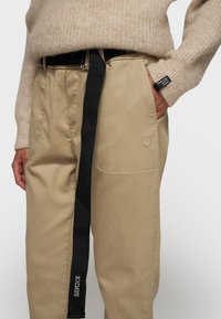 Beige katoenen broek met een zwarte webbandriem, met een zichtbare zak en het reliëfnummer "10" aan de zijkant. Zachte textuur, casual ontwerp.