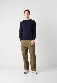 Modra bombažna pulover s dolgimi rokavi in majhnim logotipom na prsih, v kombinaciji s bež chinos hlačami in svetlimi supergami.