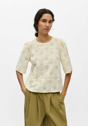 Femme aux cheveux foncés en chignon bas portant un chemisier en dentelle crème à manches bouffantes et un pantalon ample vert olive, les mains dans les poches.